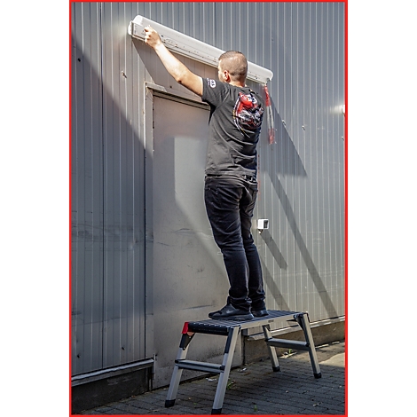 Ein Mann installiert eine Markise über einer Tür; er steht auf einer Trittleiter. Die Tür ist in eine graue Metallwand eingelassen.